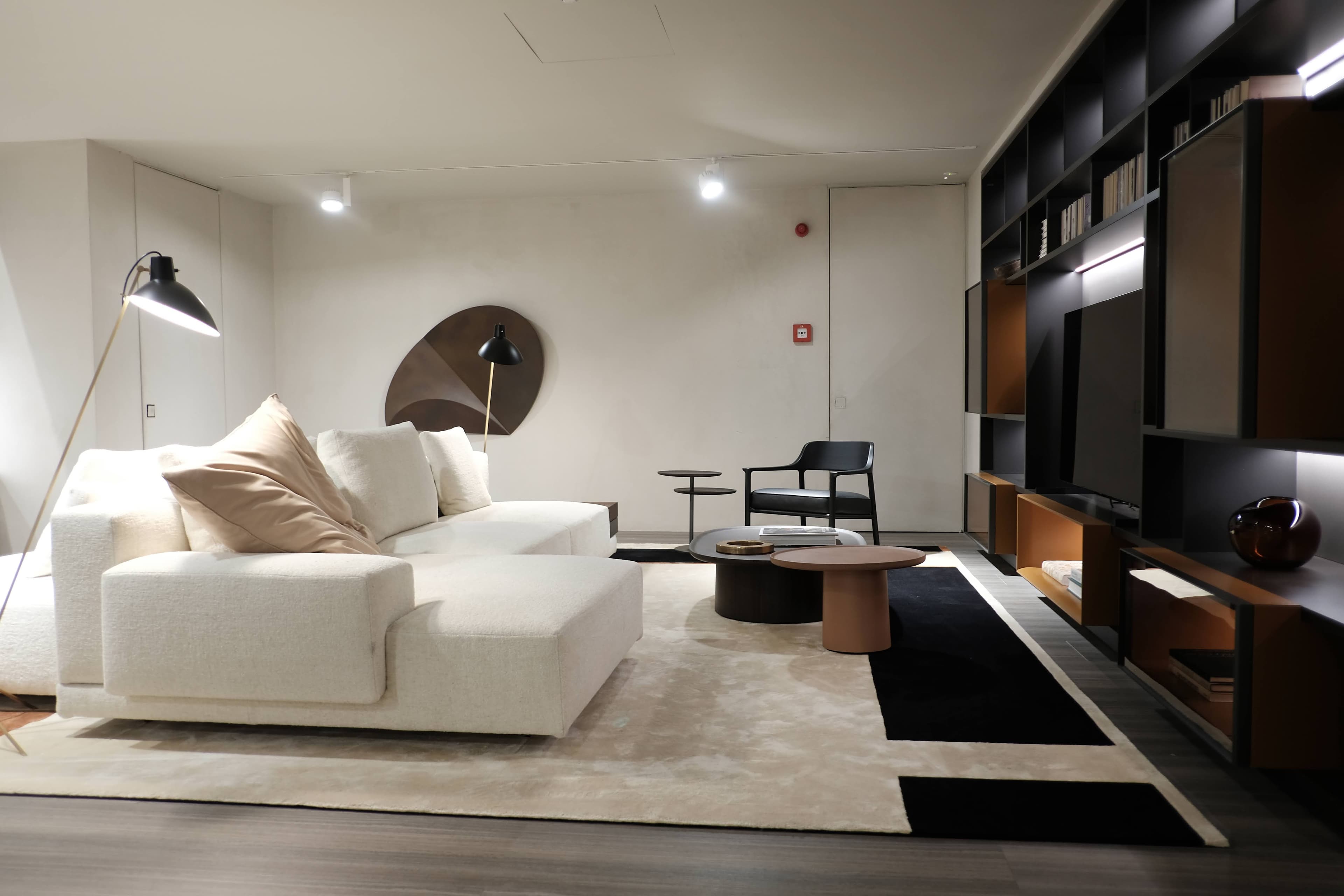 A modern and luxurious living room showroom featuring a large cream sectional sofa, a collection of round coffee tables, a black armchair, and a sophisticated dark wood shelving unit with integrated lighting. The space is adorned with a large abstract wall art piece and a contemporary floor lamp, creating an inviting and elegant atmosphere.