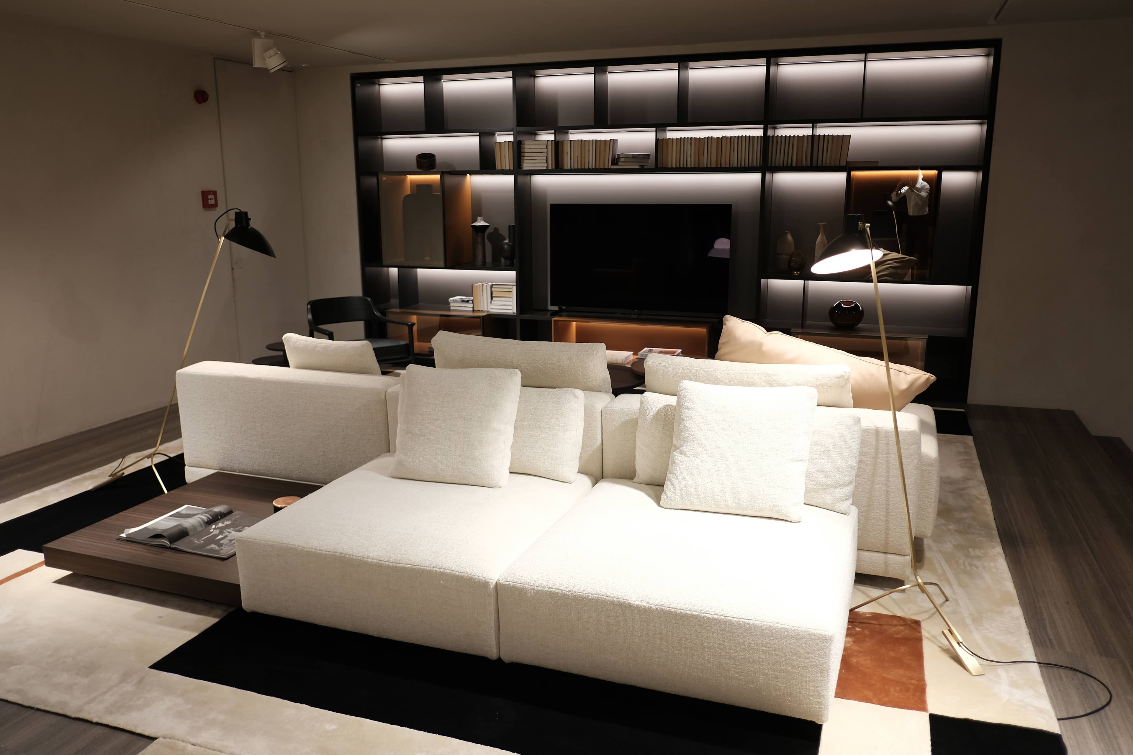 A modern and luxurious living room setup in a showroom, featuring a large, off-white modular sectional sofa, a dark wood coffee table with a magazine, and a sophisticated dark shelving unit with integrated lighting and a television. Two elegant floor lamps illuminate the space, which is anchored by a geometric area rug.