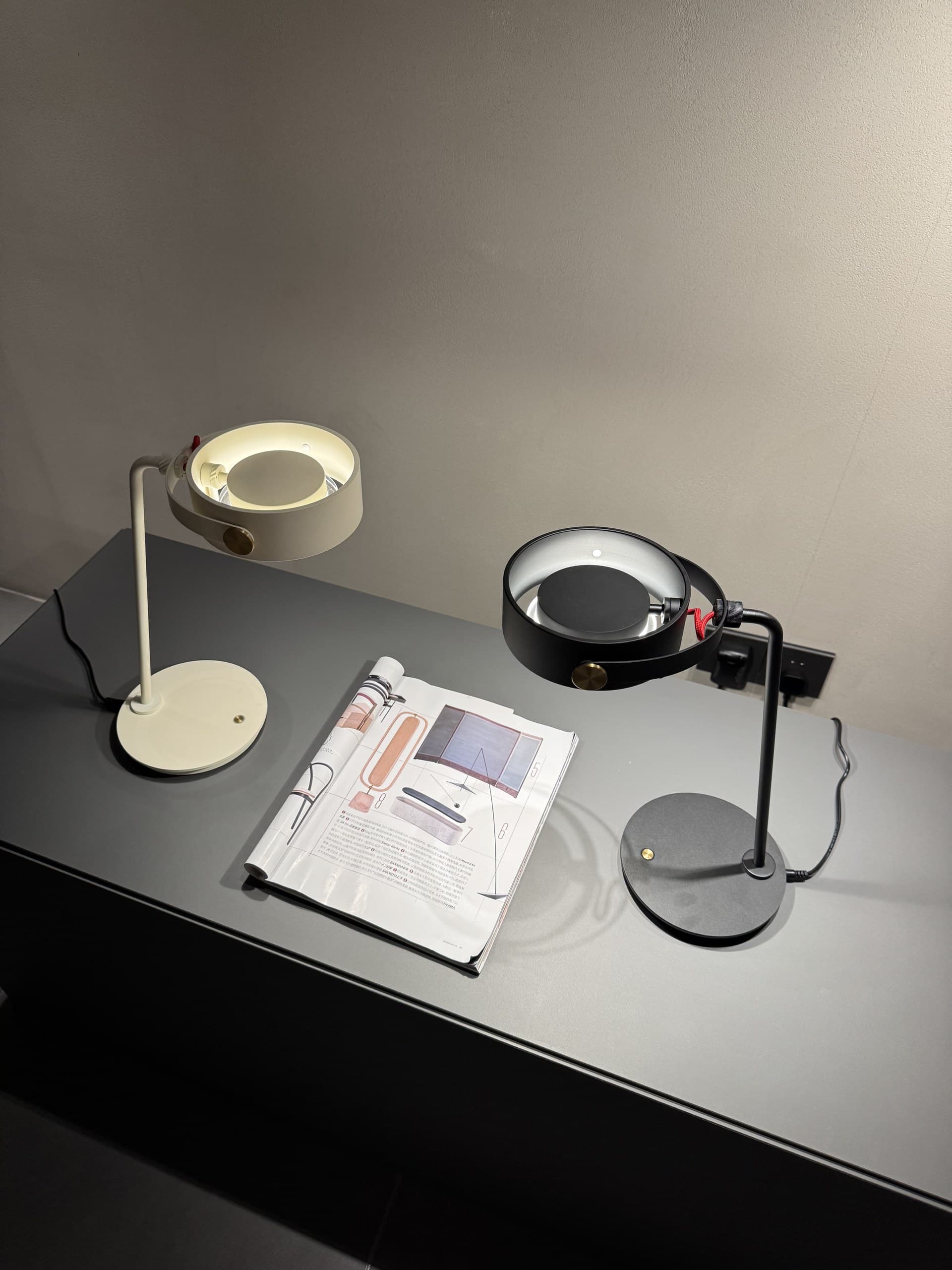 Two modern desk lamps, one white and one black, illuminate a magazine on a dark gray surface.