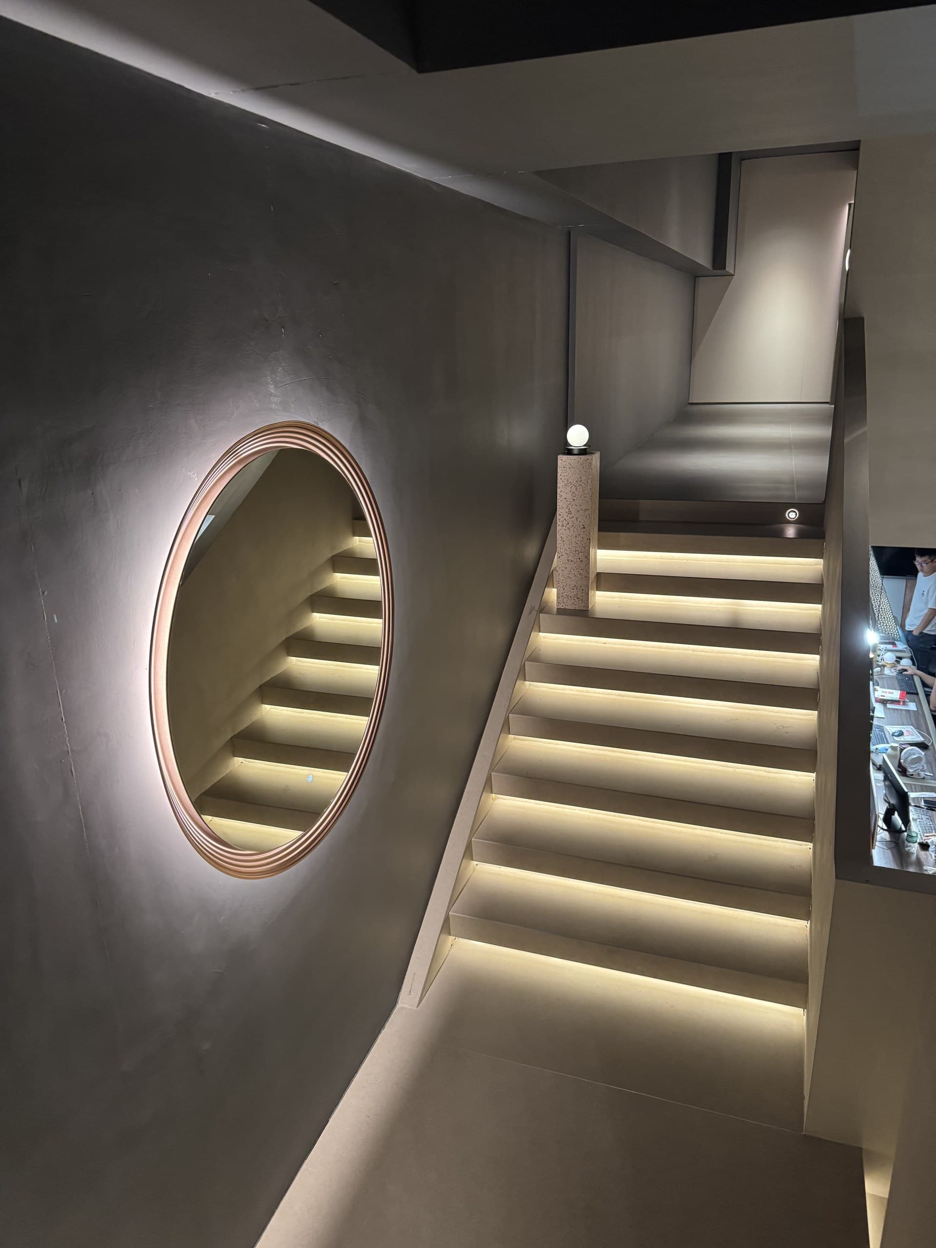 A modern staircase with illuminated steps and a round mirror reflecting the stairs, set in a dimly lit interior.