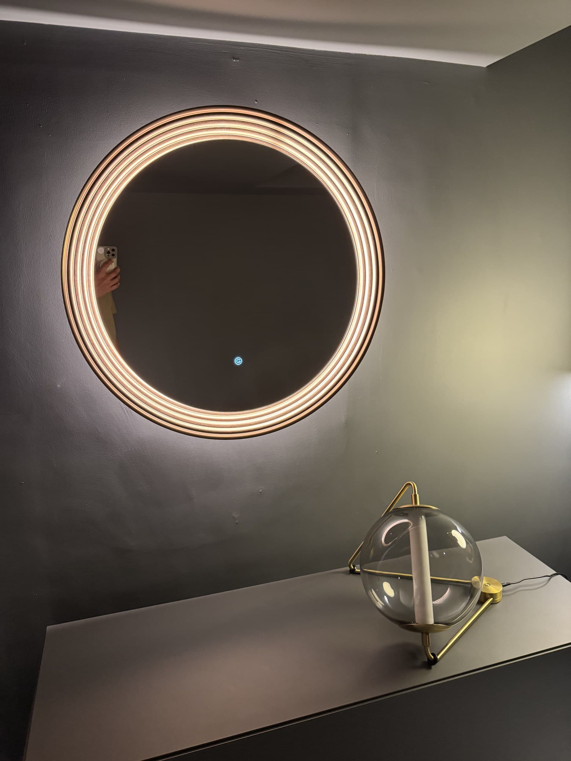 A round mirror with an illuminated frame hangs above a console table, next to a spherical table lamp.