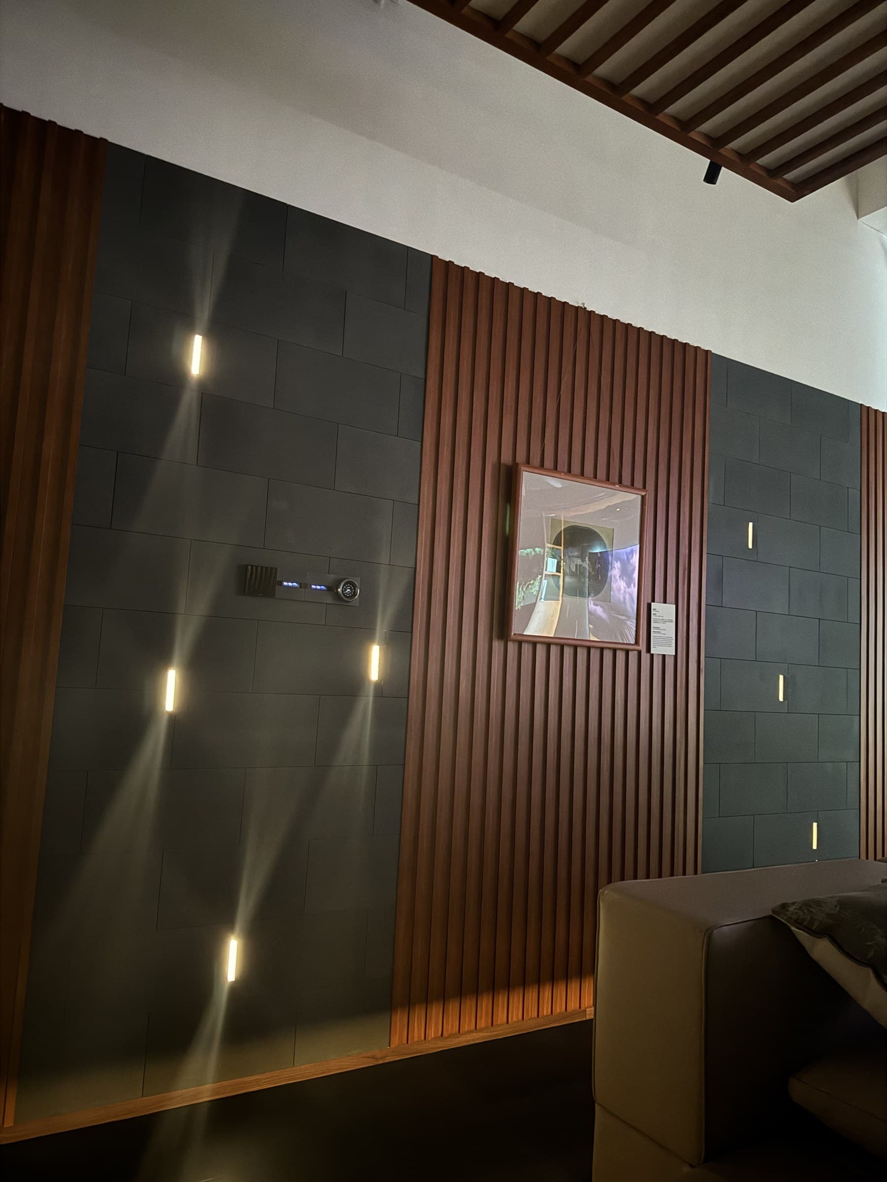 A modern wall design with alternating dark grey panels and wooden slats, featuring several glowing wall lights and a smart home control panel. A framed picture hangs on the wooden section, and a light-colored sofa is partially visible.