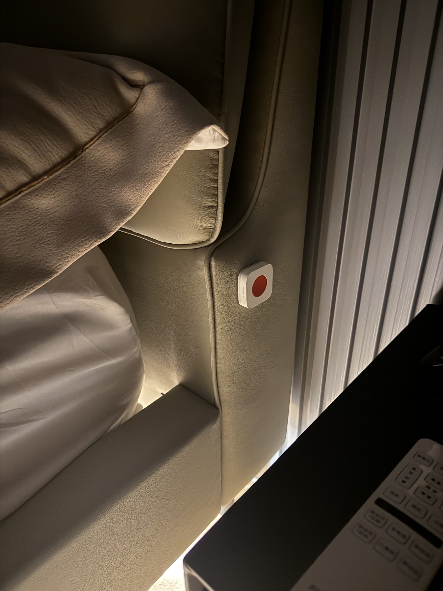 A white smart button with a red center is attached to a light brown headboard in a bedroom setting.