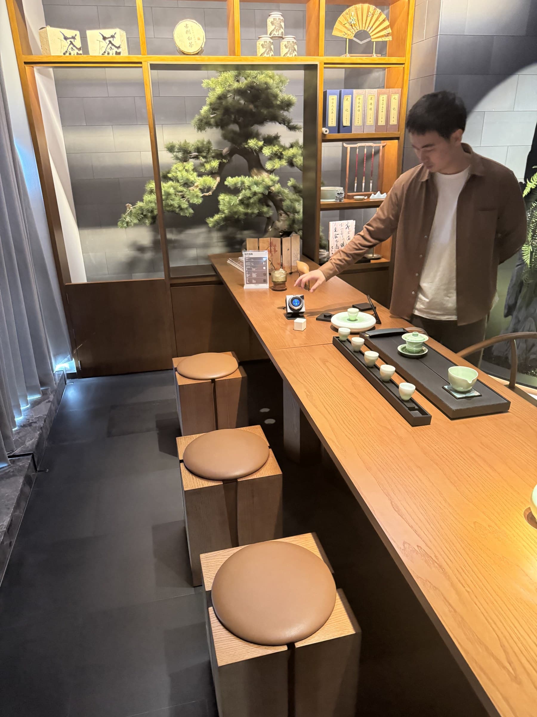 A man in a brown shirt and white t-shirt stands at a long wooden table, reaching for a smart home device. The table is set with multiple tea cups and a tea pot. In the background, there's a display with a large artificial bonsai tree and shelves holding various decorative items like books, a fan, and tea containers. Three wooden stools with round brown cushions are visible in the foreground.