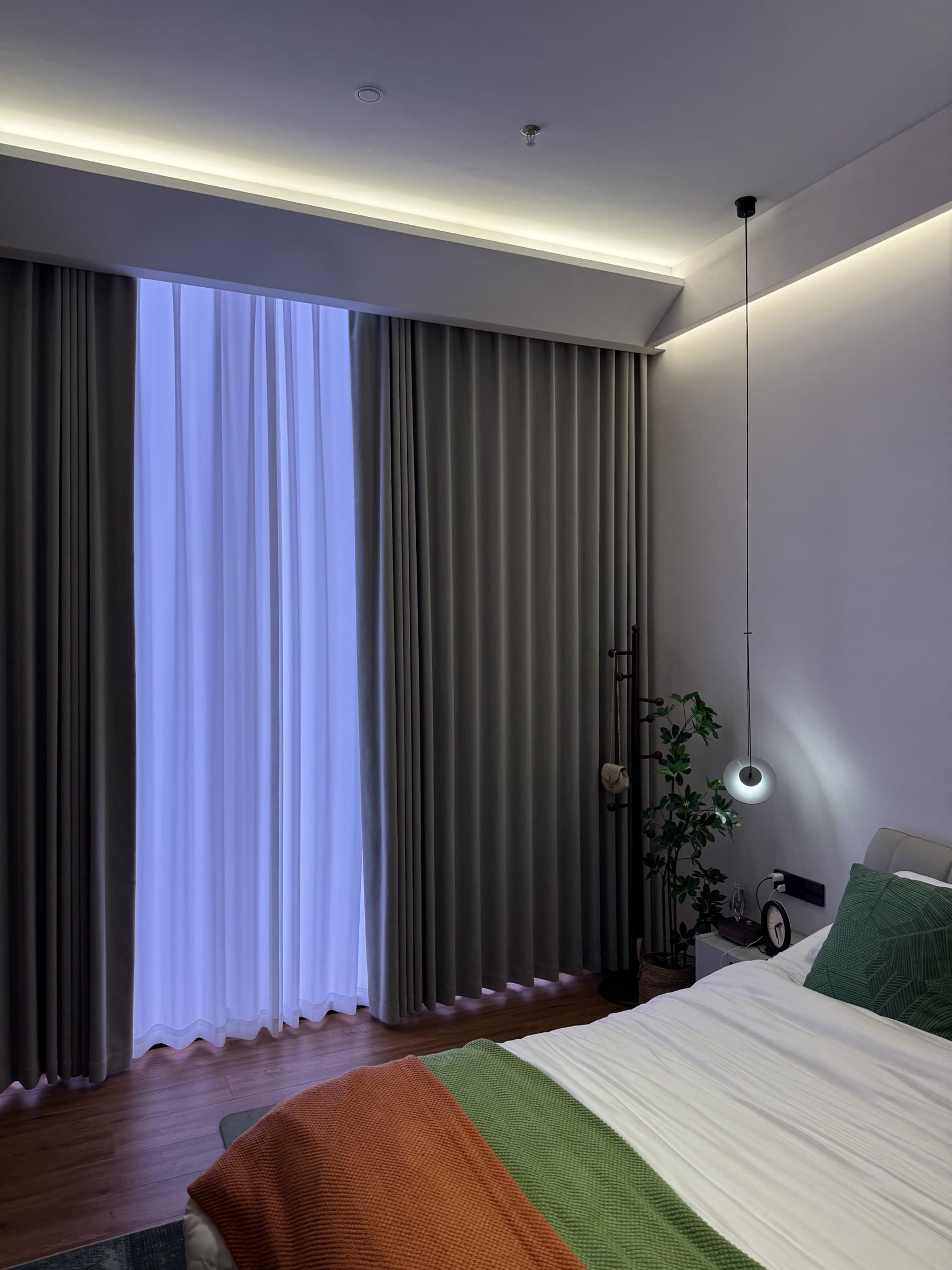 A modern bedroom with a bed, bedside table, plant, and hanging light, illuminated by cove lighting and natural light from a window with curtains.