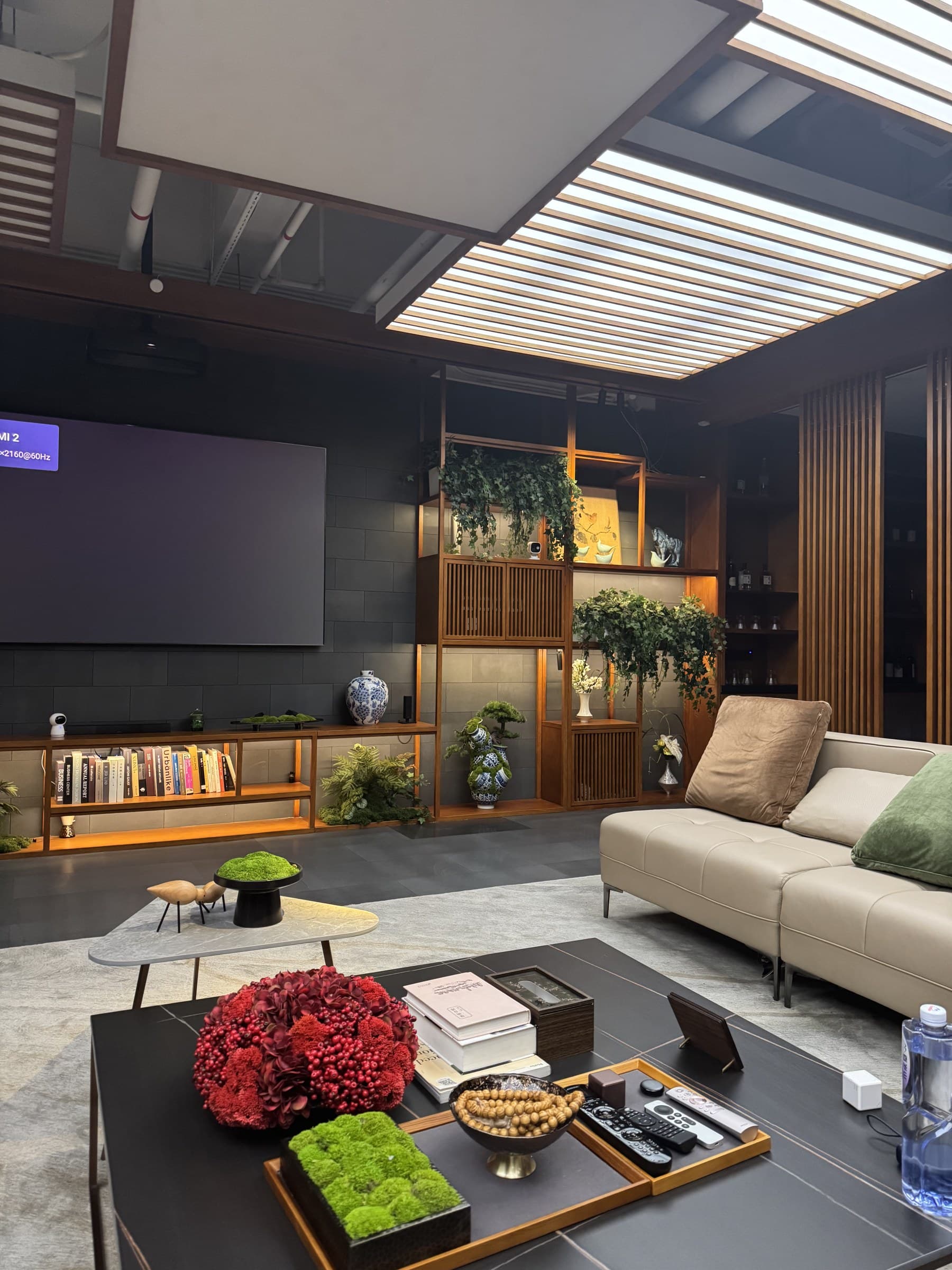A stylish living room featuring a large TV, a comfortable beige sofa, and a dark coffee table with various decorations. Wooden shelves filled with plants and ornaments line the back wall.