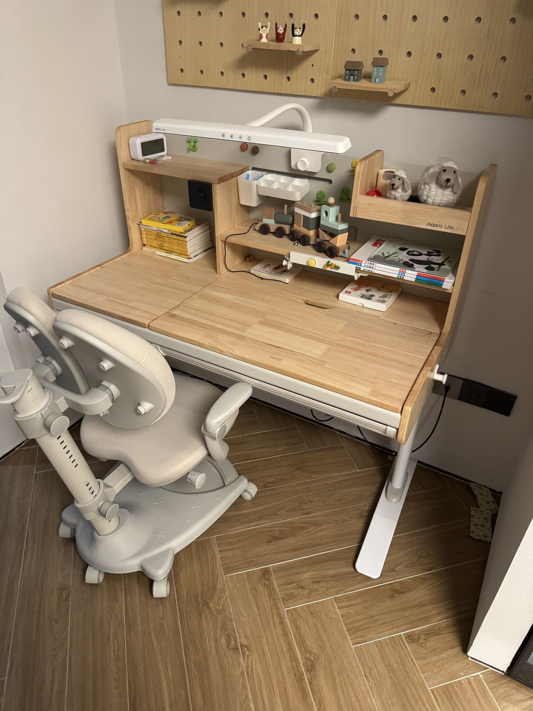 A light-colored wooden desk with a white adjustable chair. The desk has shelves with books, small decorative houses, and stuffed sheep. A white desk lamp is mounted above the desk, and a wooden pegboard is on the wall above.