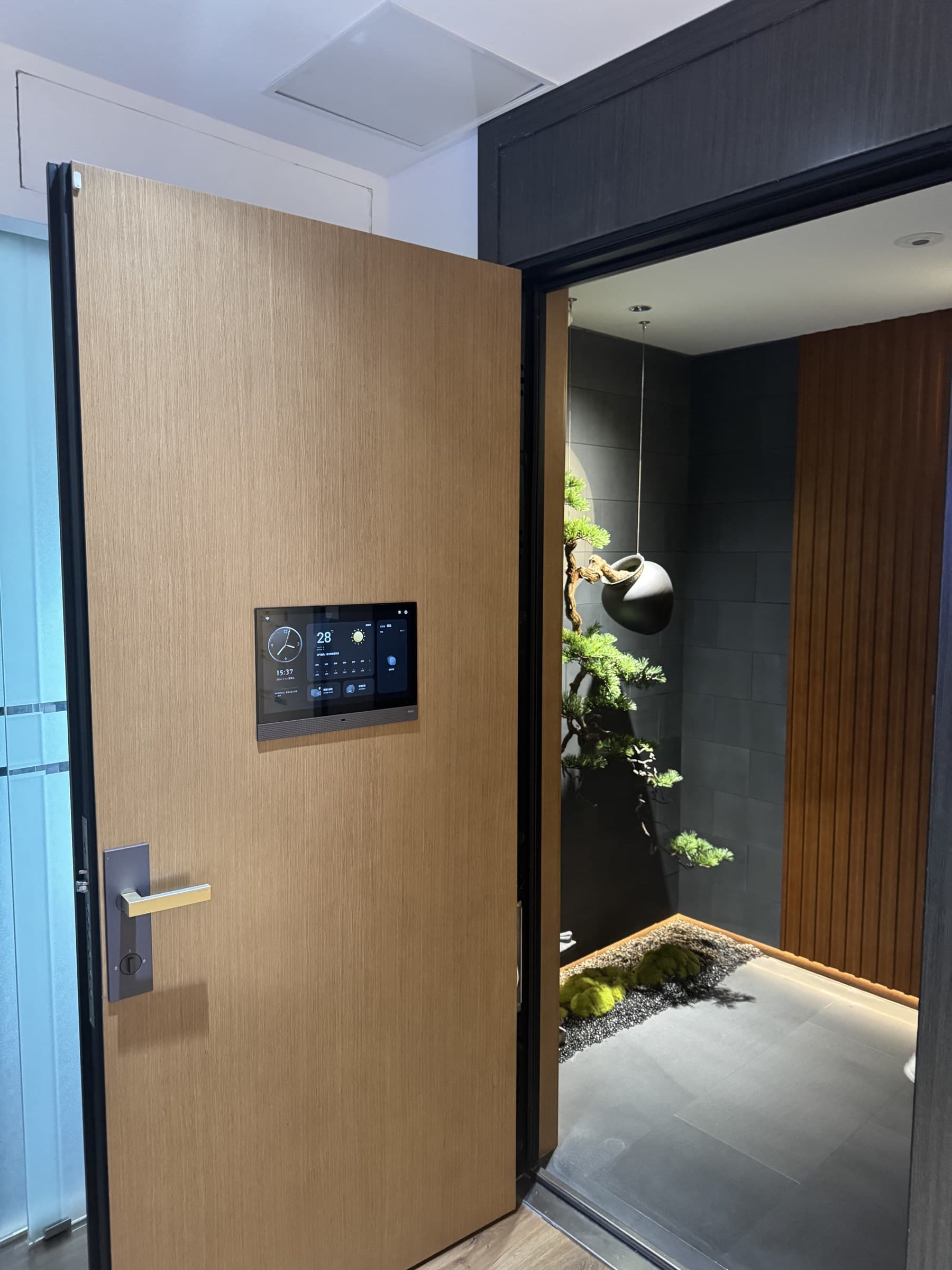 An open wooden door with a smart panel and a modern handle. The panel displays time, weather, and smart home controls. Beyond the door, a decorative space with dark tiled walls, a wooden slatted wall, and a hanging pot with greenery is visible.