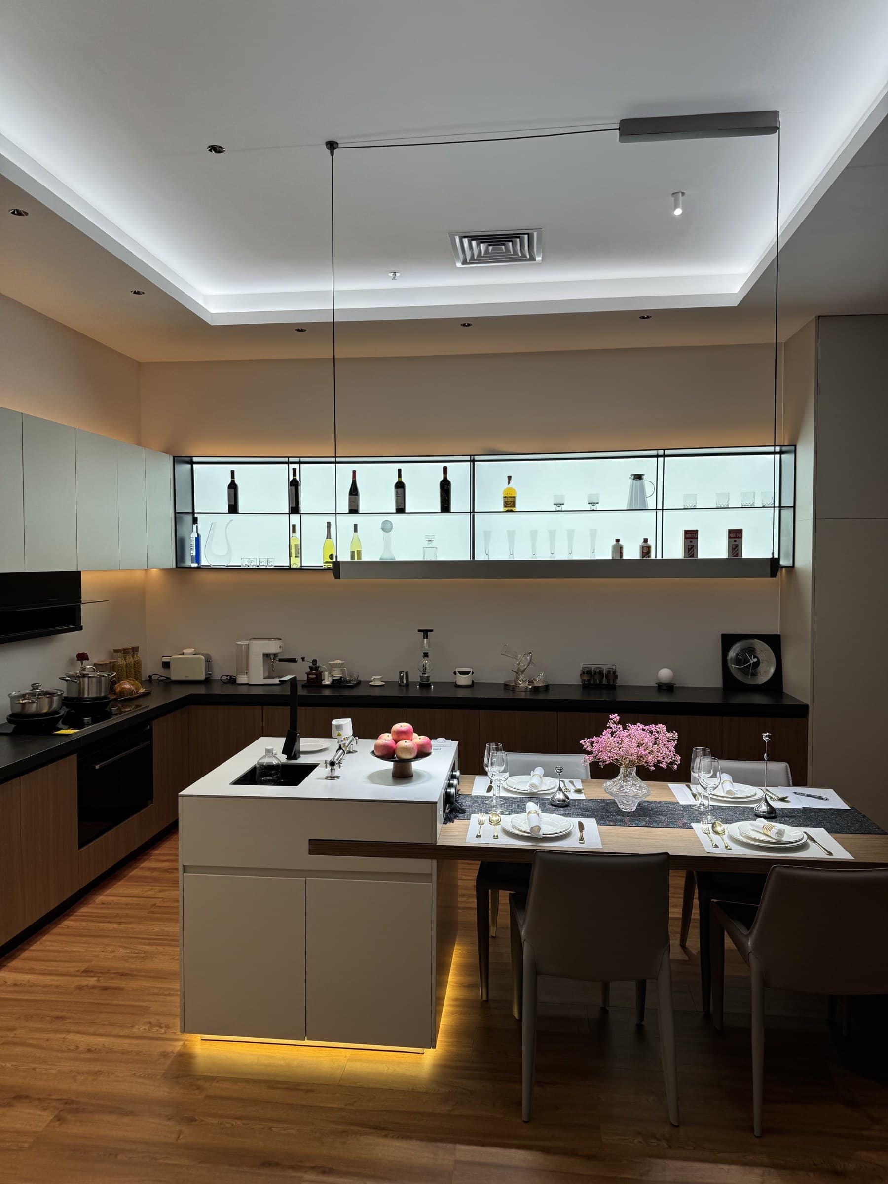 A modern kitchen and dining area featuring a kitchen island with a sink, a dining table set with place settings, and a display shelf with bottles and glassware. The room has recessed lighting and under-cabinet lighting.