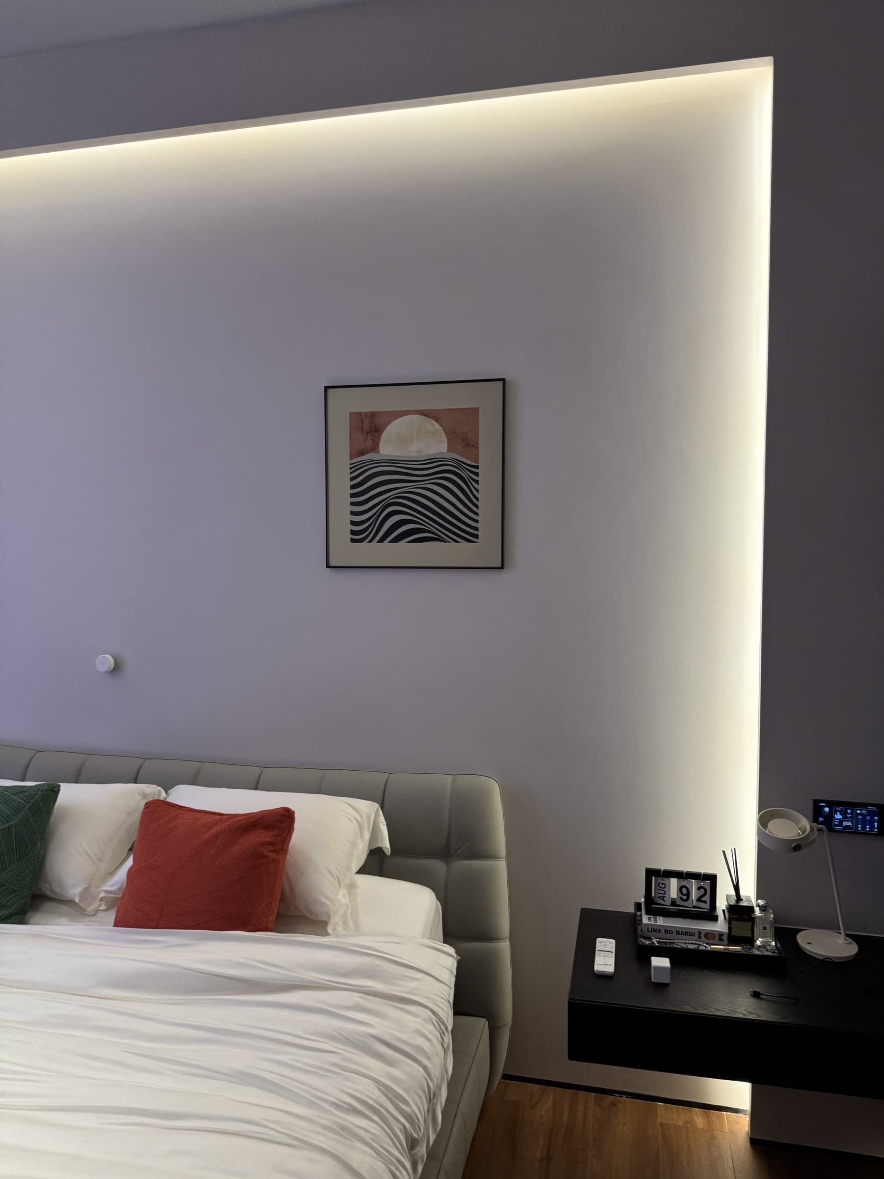 A modern bedroom with a bed, nightstand, and artwork, illuminated by hidden strip lighting.
