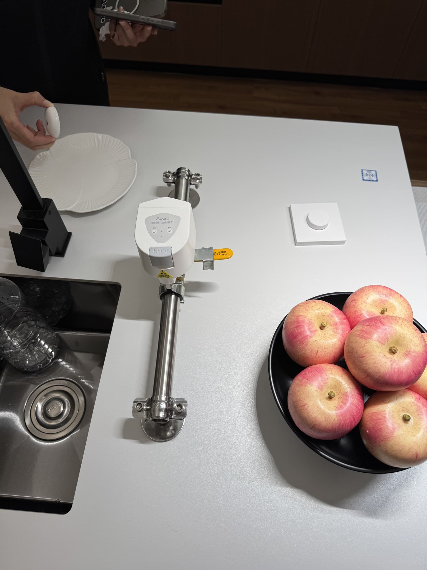 A person's hands are visible in a kitchen setting, with one hand holding a white device and the other holding a smartphone. An Aqara smart valve controller is mounted on a pipe next to a sink, and a bowl of apples sits on the counter.