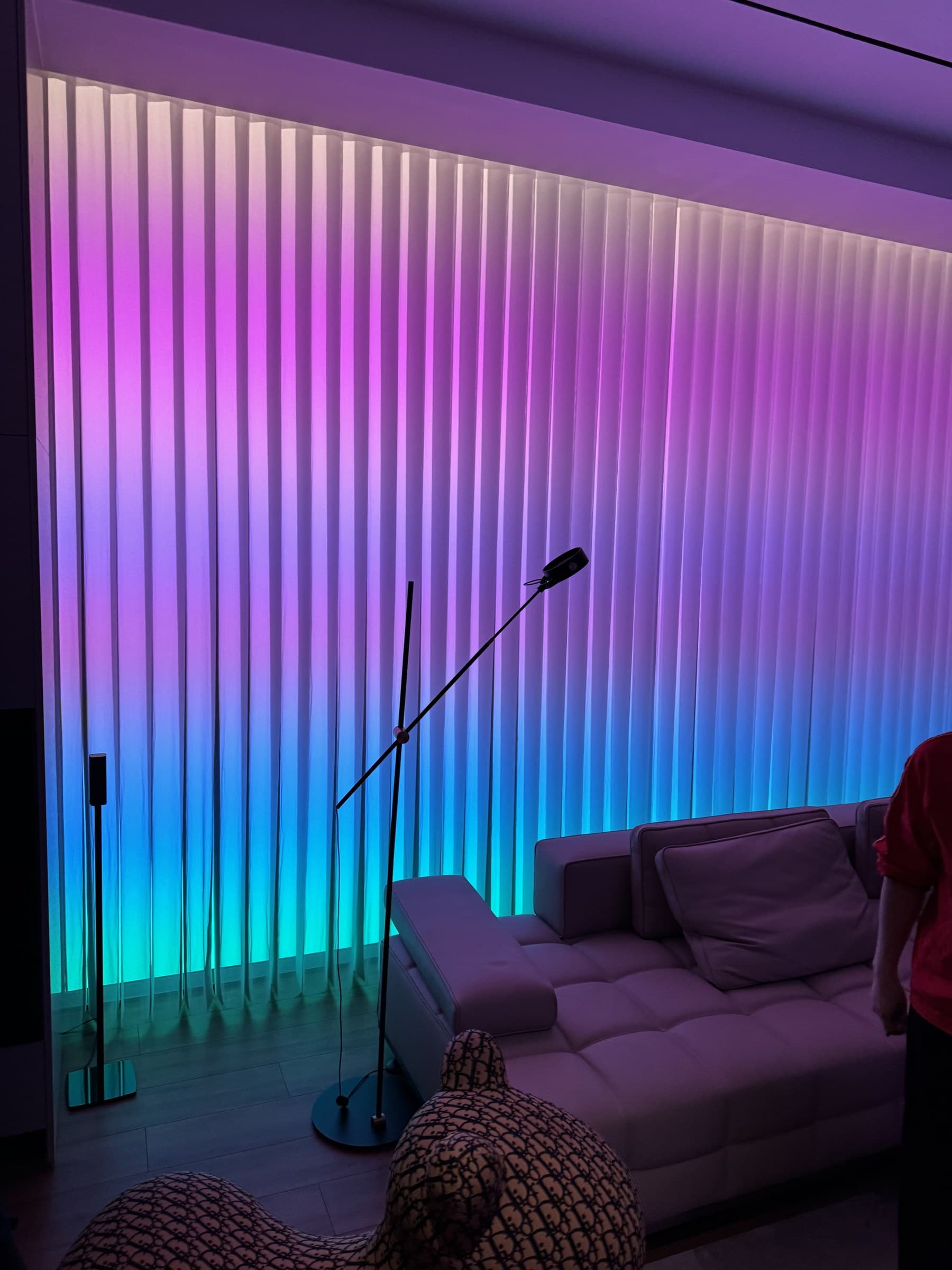 A modern living room with vertical blinds illuminated by a colorful gradient of purple, blue, and green light. A light-colored sofa and a floor lamp are also visible.