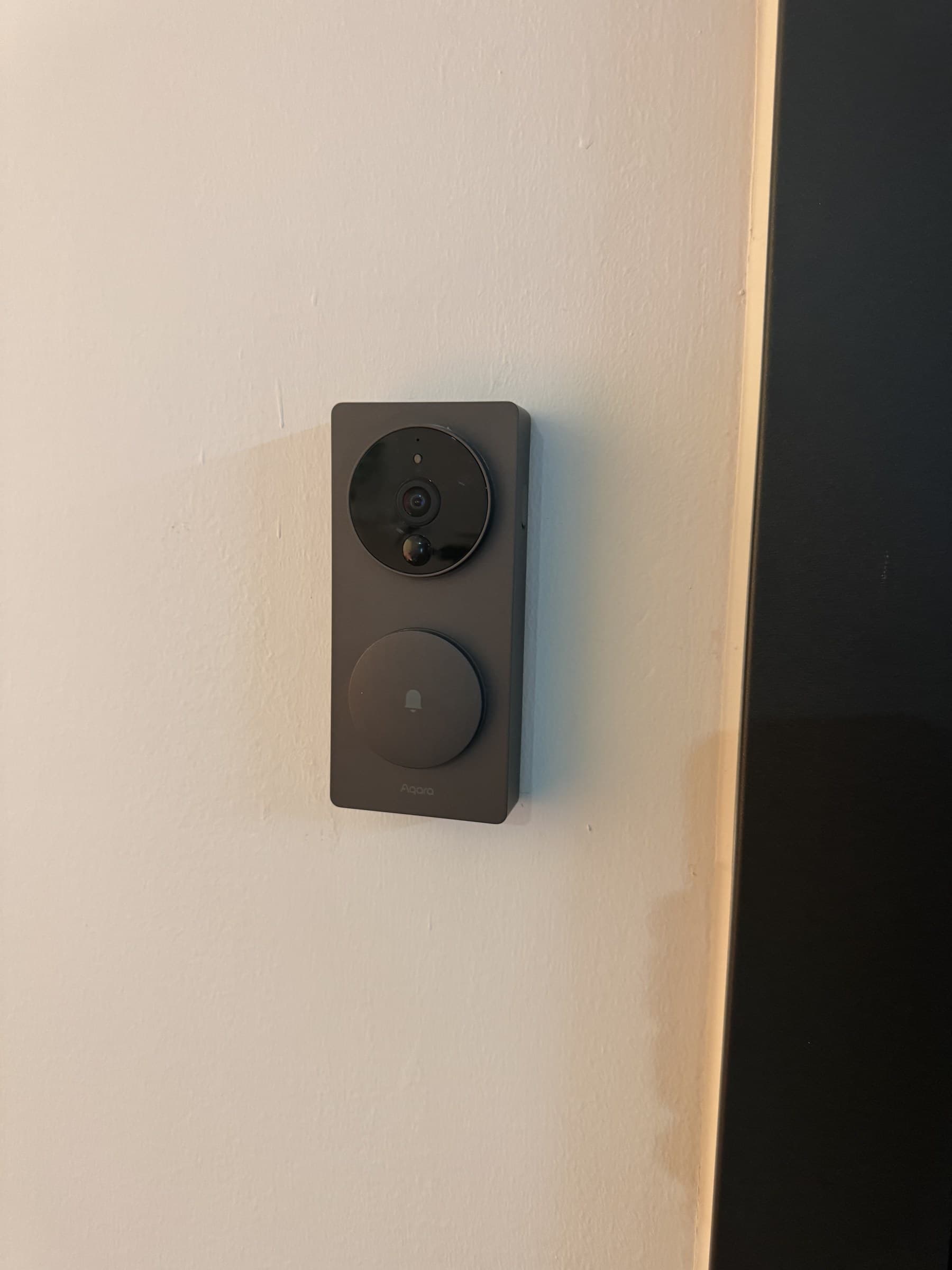 A dark gray Aqara video doorbell with a camera lens and a circular doorbell button with a bell icon is mounted on a light-colored wall.