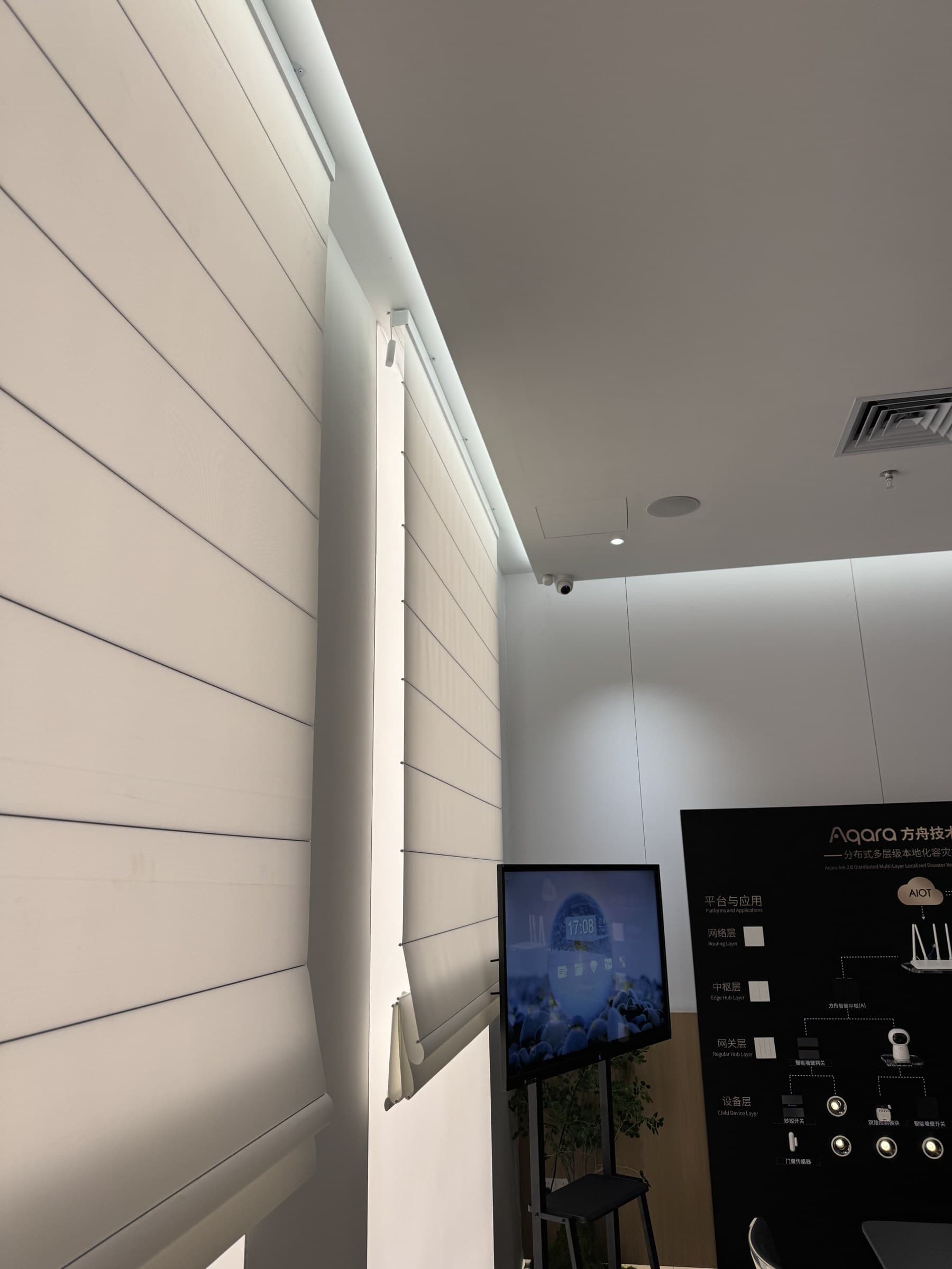 A showroom with white blinds, a large screen displaying a digital clock and a smart home system diagram, and ceiling-mounted cameras.